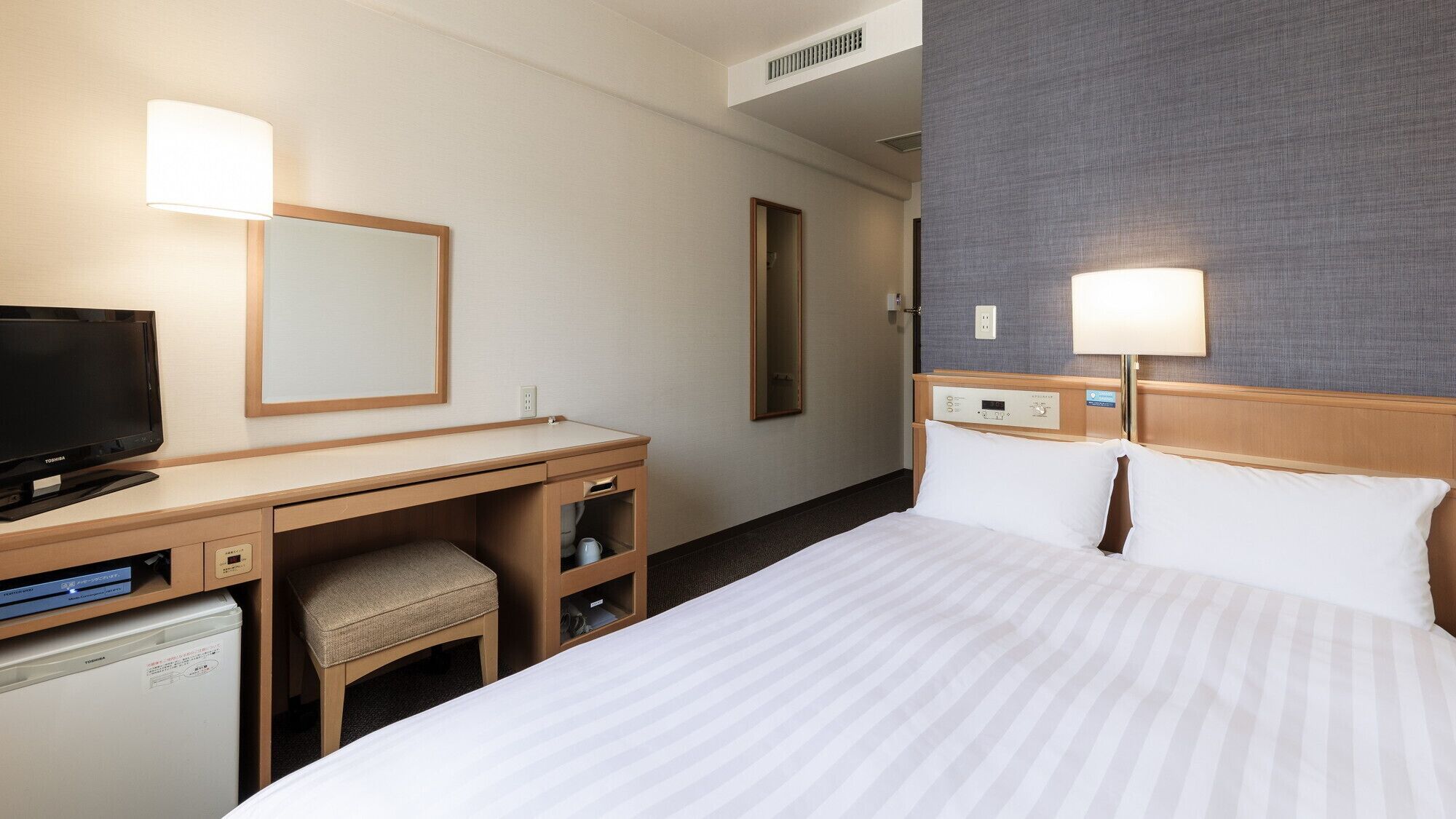 A hotel room with a bed, a desk with a TV, a chair, a mirror, and a wall-mounted lamp.