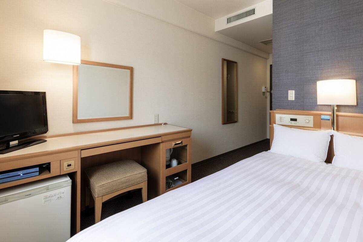 A hotel room with a bed, a desk with a TV, a chair, a mirror, and a wall-mounted lamp.