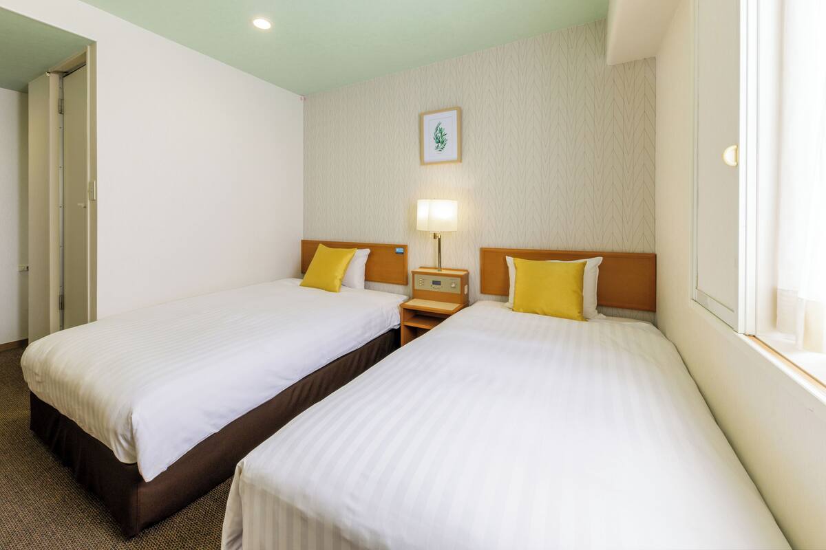 A hotel room with two single beds, a nightstand with a lamp, a framed picture on the wall, and a window with curtains.