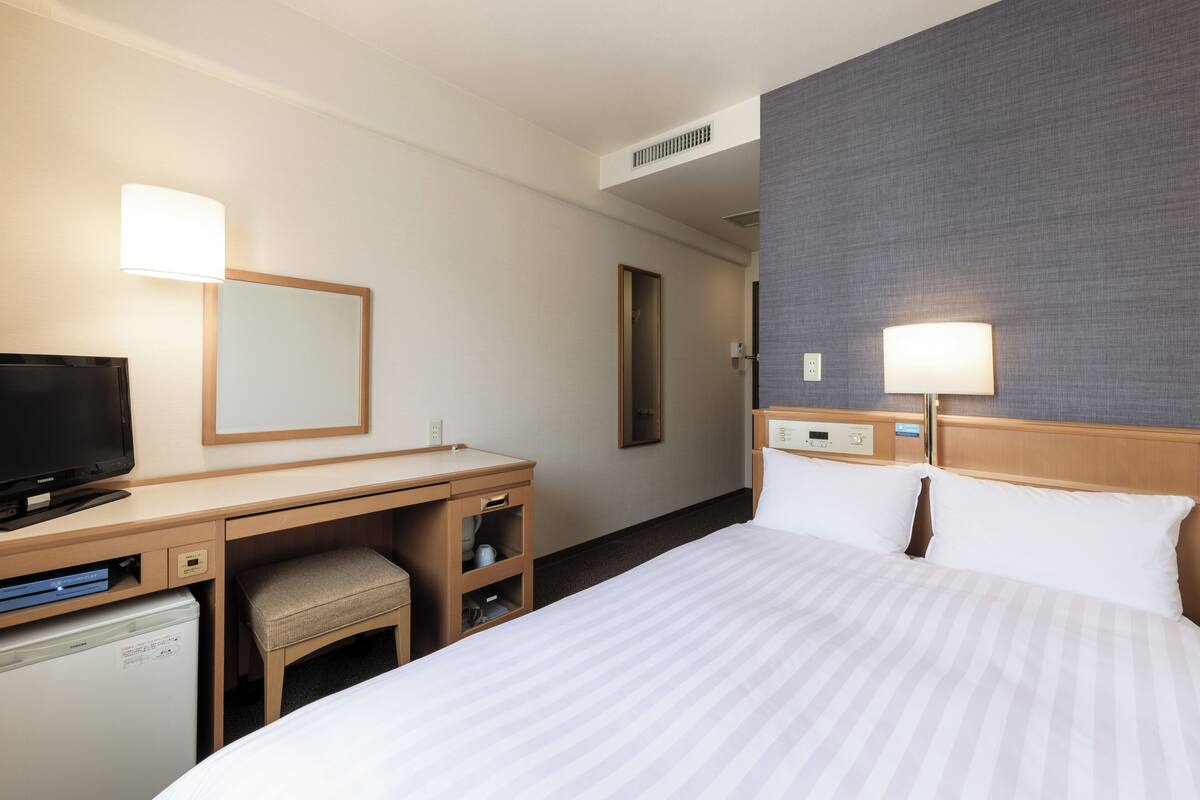 A hotel room with a bed, a desk with a television, a chair, a small refrigerator, and a mirror.