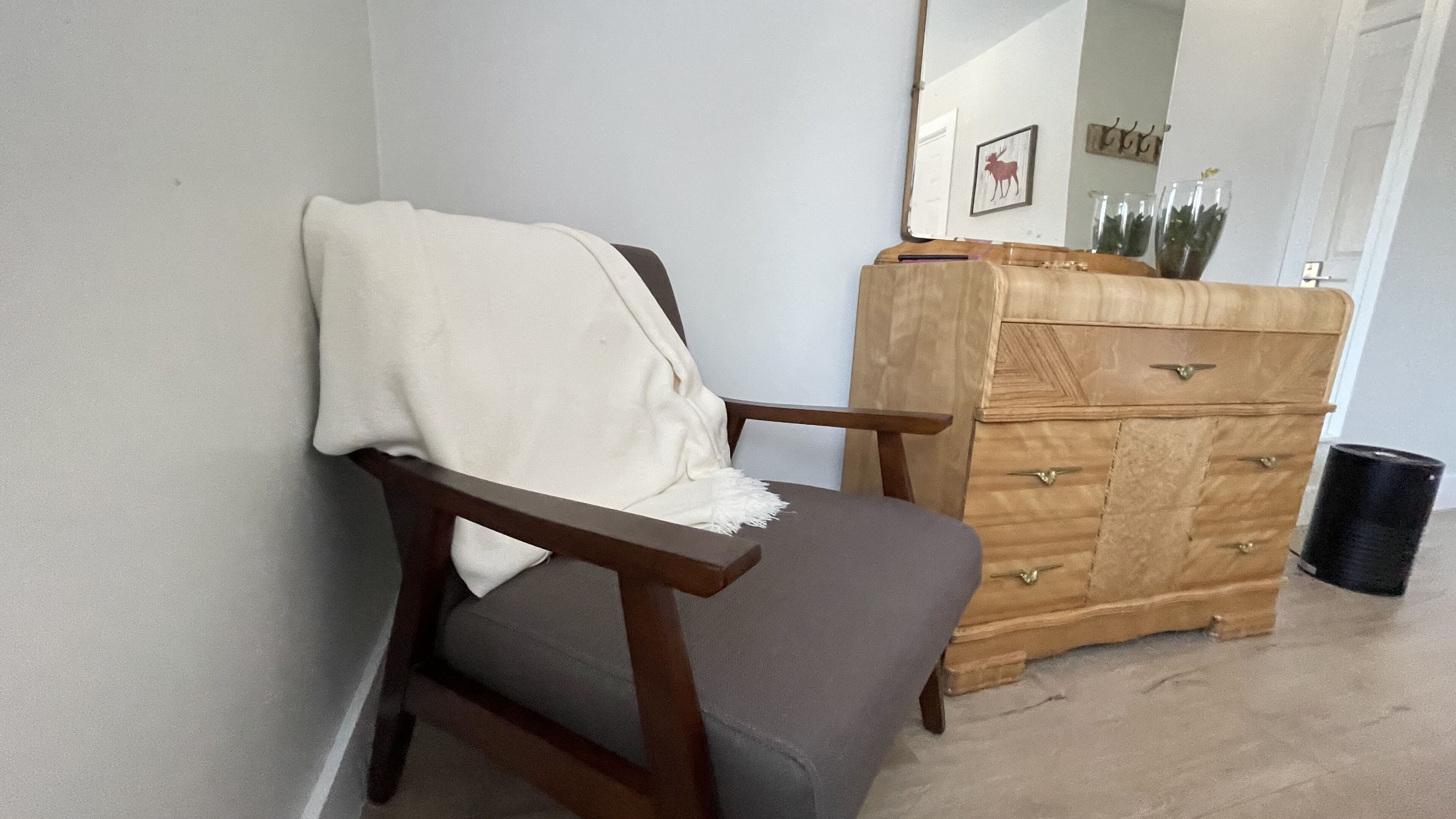 Una silla con una manta blanca, un aparador de madera con cajones y un cubo de basura.