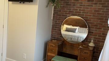 A bedroom with a brick wall, a television mounted above a dresser, a hanging plant, and a bed with patterned bedding.