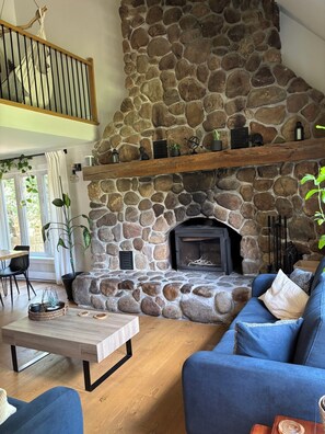 Una chimenea de piedra con repisa de madera, un sofá azul y un comedor con mesa y sillas de madera.