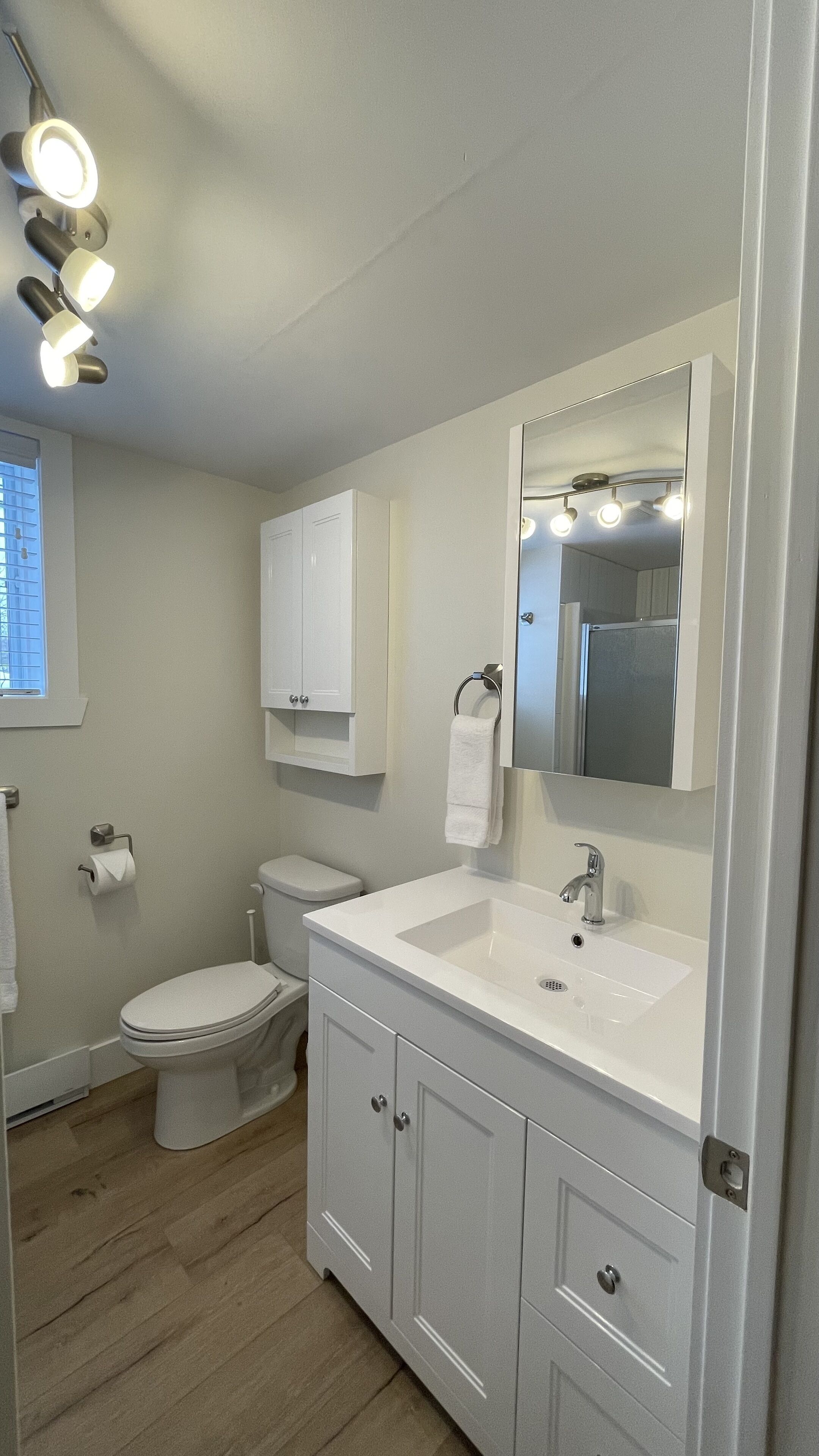 Une salle de bain moderne avec un lavabo blanc, un meuble-lavabo et des toilettes. Un miroir avec des lumières est placé au-dessus du lavabo, et il y a une fenêtre avec des stores.