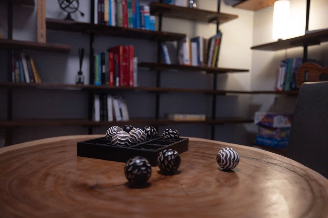 Une table ronde en bois, ornée de sphères décoratives, et un plateau noir, dans une pièce dont le fond est constitué d’étagères à livres.