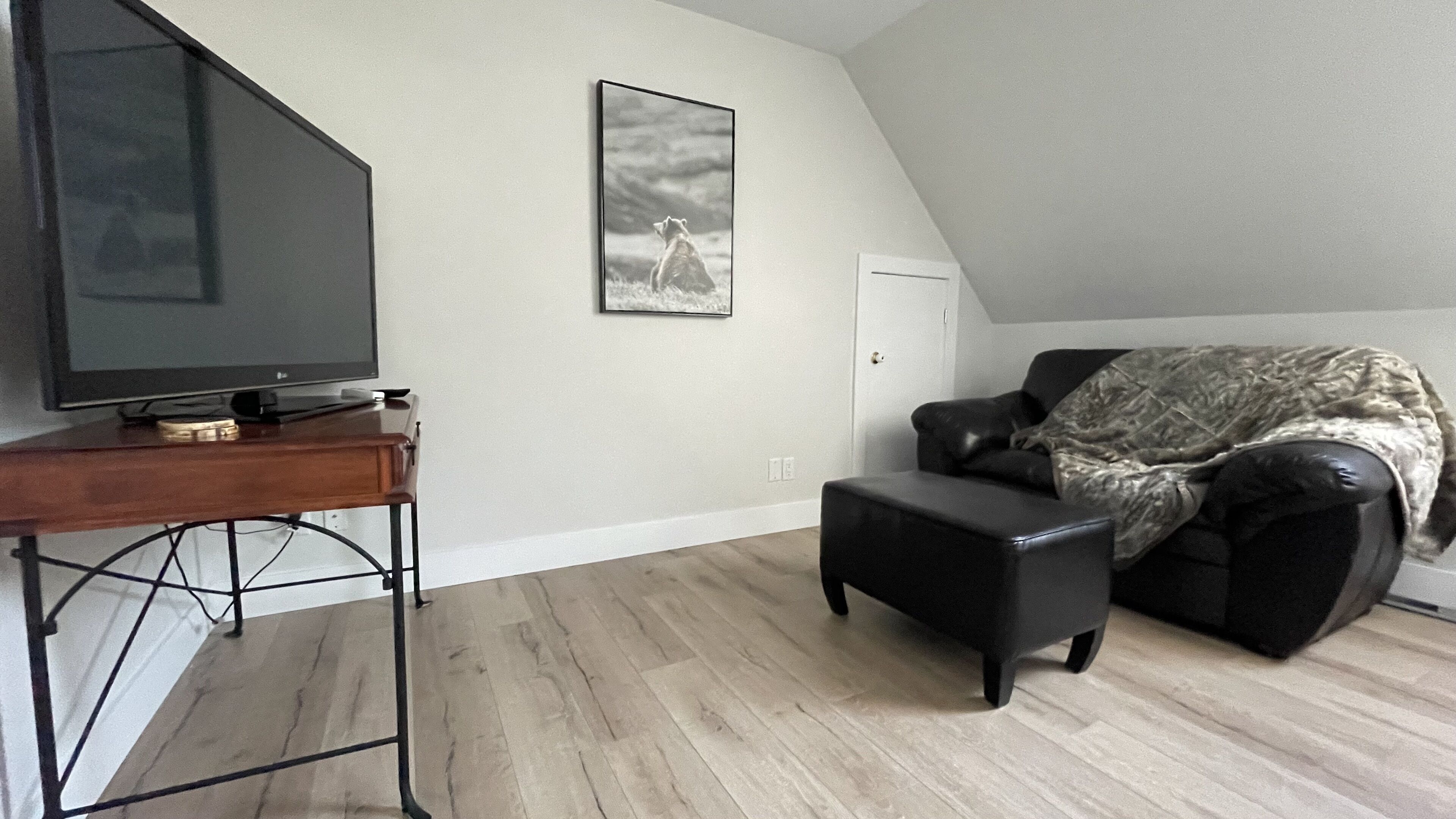 Una sala de estar con una televisión de pantalla plana sobre una mesa de madera, un sofá de cuero negro, un cuadro en la pared y una puerta.