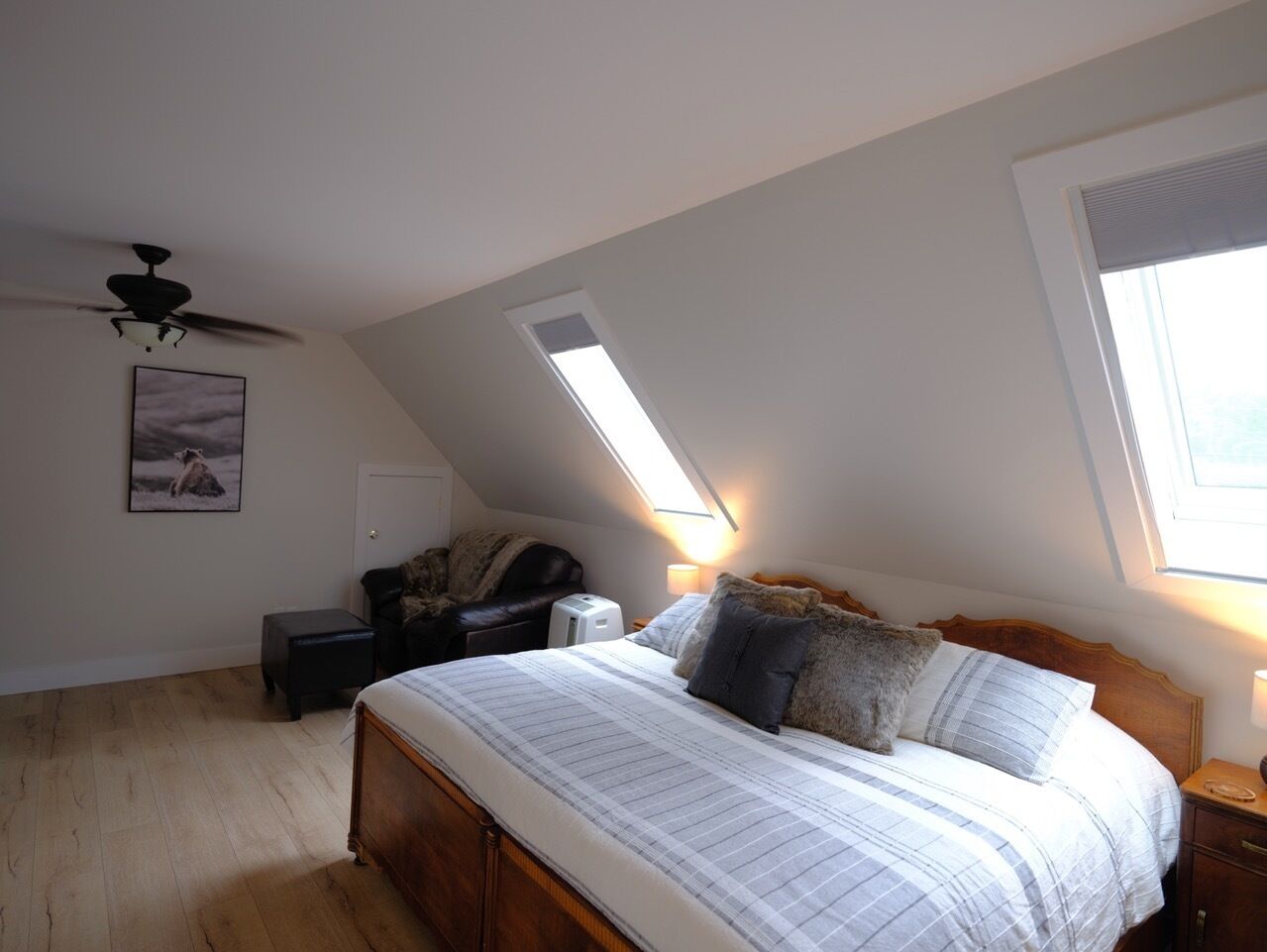 A bedroom with a sloped ceiling, a bed with a wooden headboard, a ceiling fan, a small bedside table, a nightstand, and a window with a view.