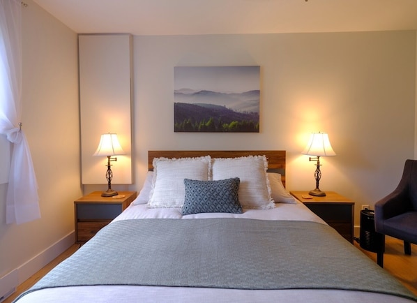 Een net opgemaakt bed met een grijze deken, twee witte kussens en een blauw gestreept kussen. Twee nachtlampjes staan op houten nachtkastjes, aan de muur hangt een schilderij van een landschap, en rechts staat een stoel.