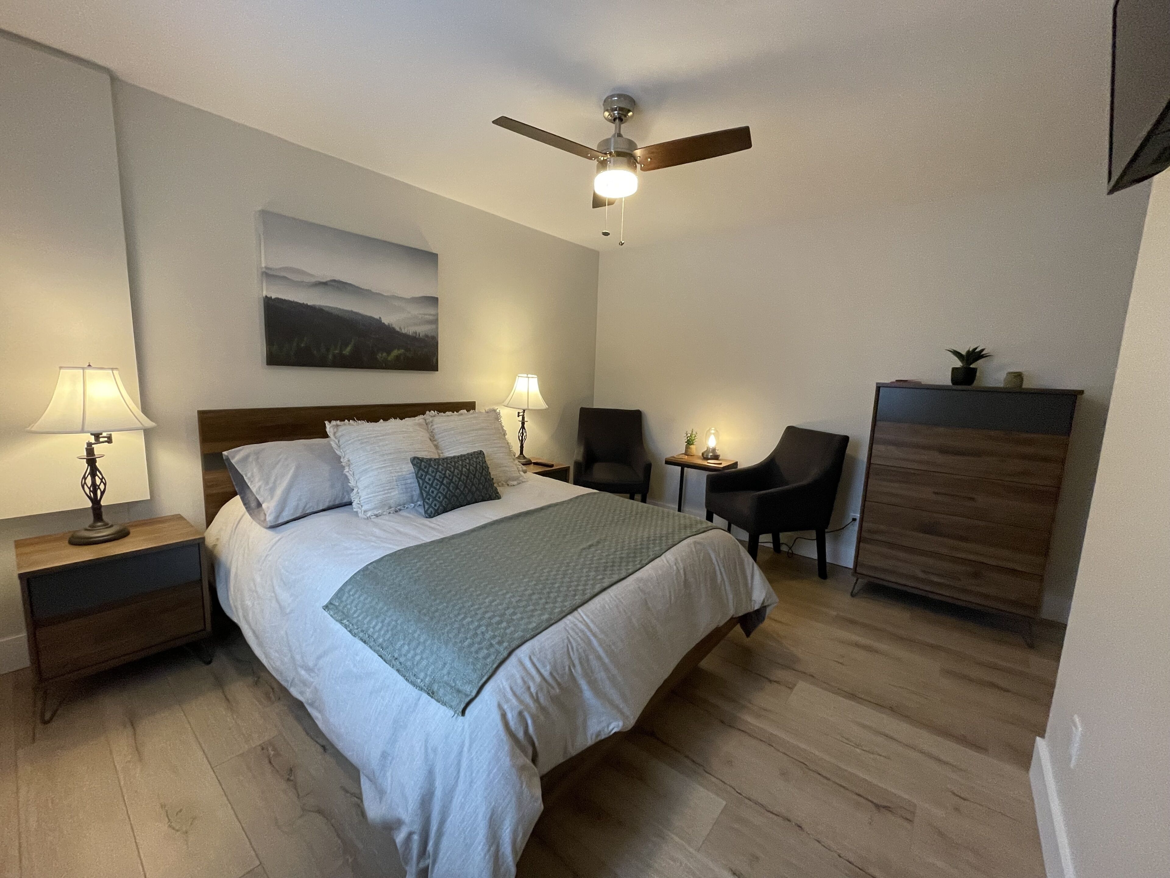 A neatly arranged bedroom with a bed, two bedside tables with lamps, a chair, a desk, and a ceiling fan.