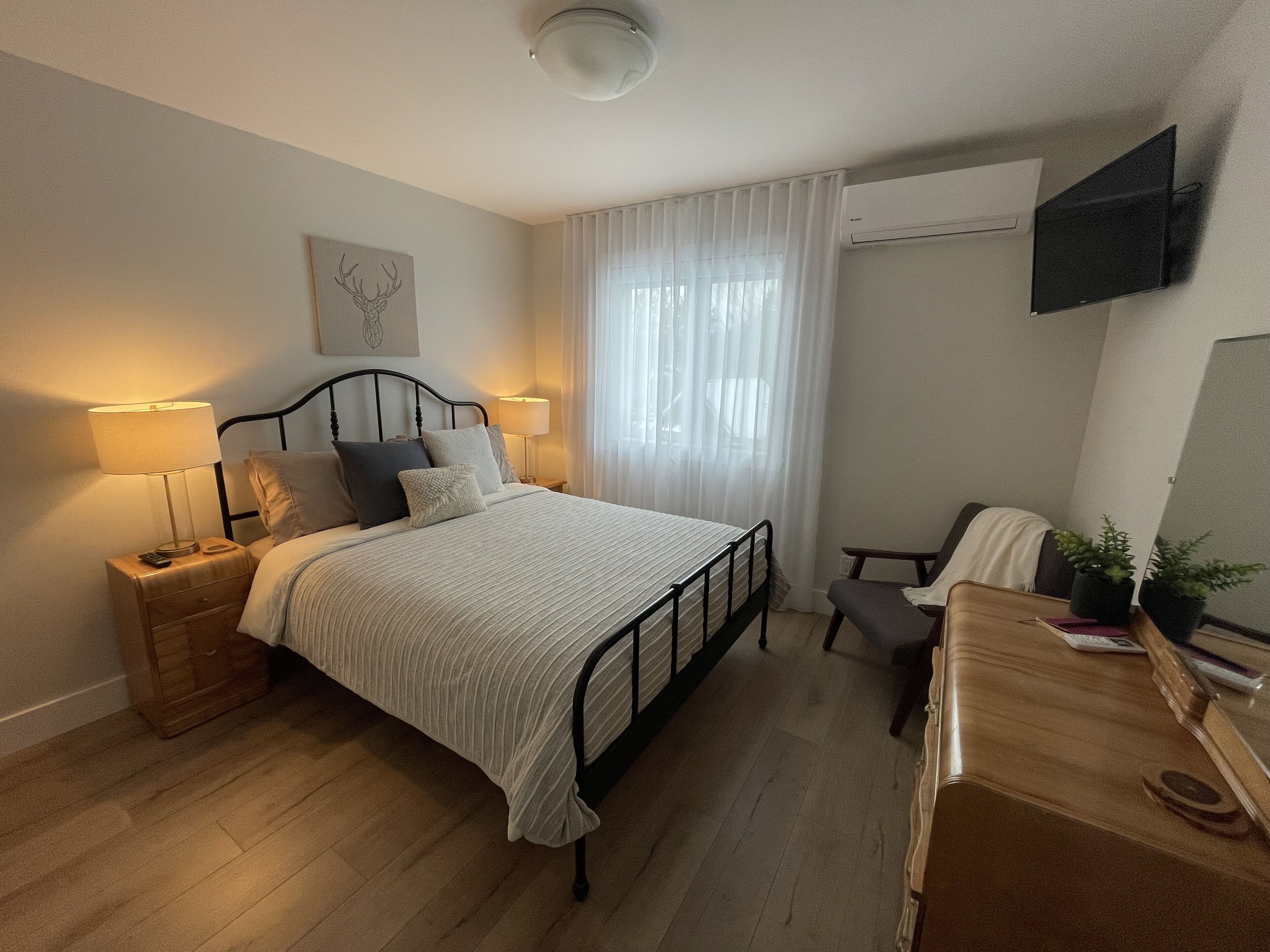 A bedroom with a bed, bedside lamps, a desk, a chair, a TV mounted on the wall, and a window with curtains.