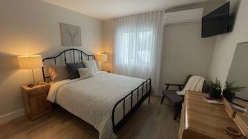 A bedroom with a bed, bedside lamps, a desk, a chair, a TV mounted on the wall, and a window with curtains.
