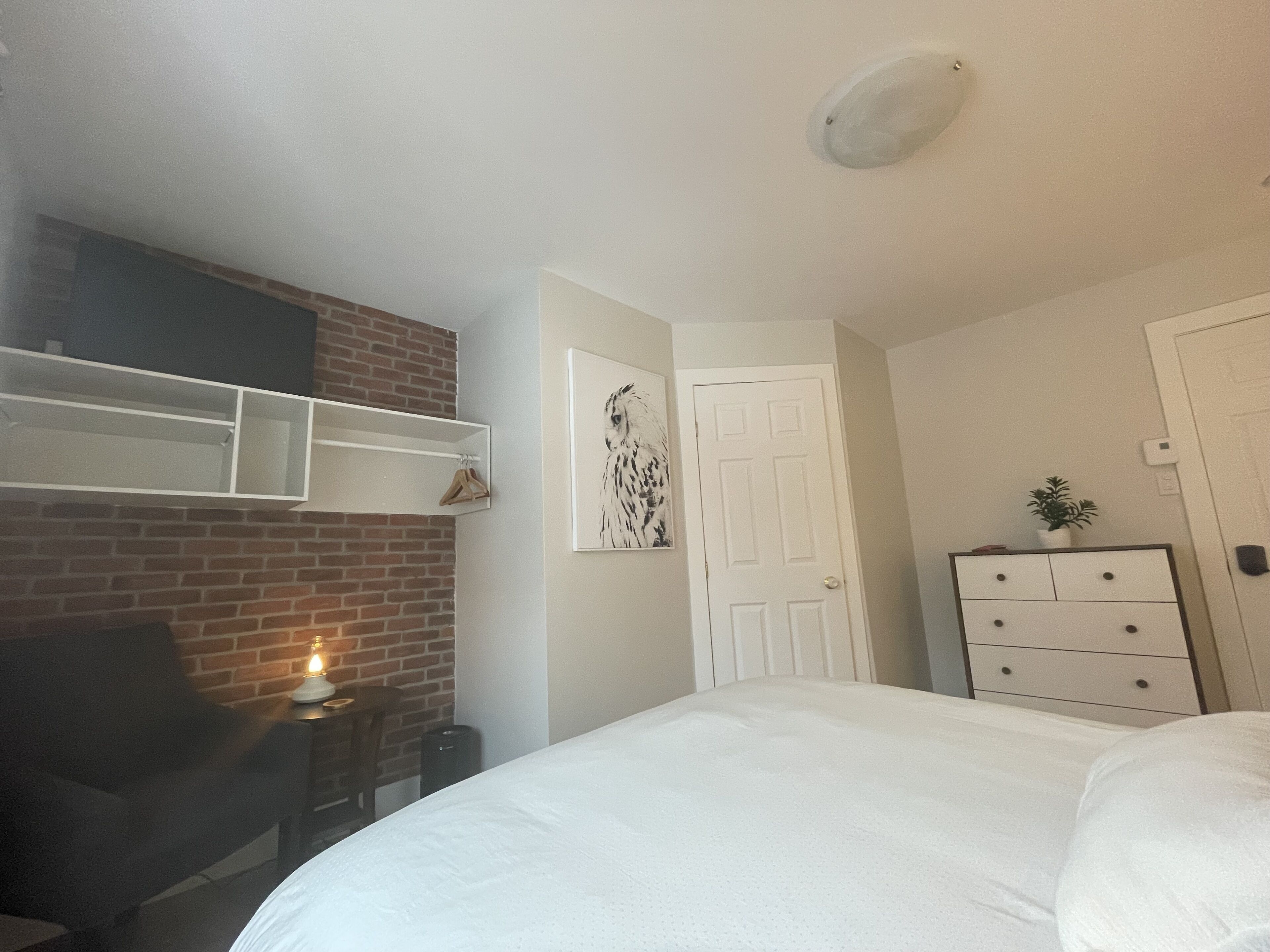 A bedroom with a brick wall, a white door, a dresser, and a framed picture on the wall.