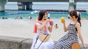 Dos mujeres sentadas en un borde de hormigón junto a una playa, con un puente al fondo.