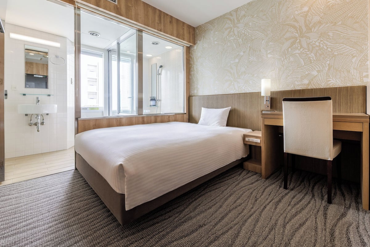 A hotel room with a large bed, a desk, a chair, a mirror, and a window.