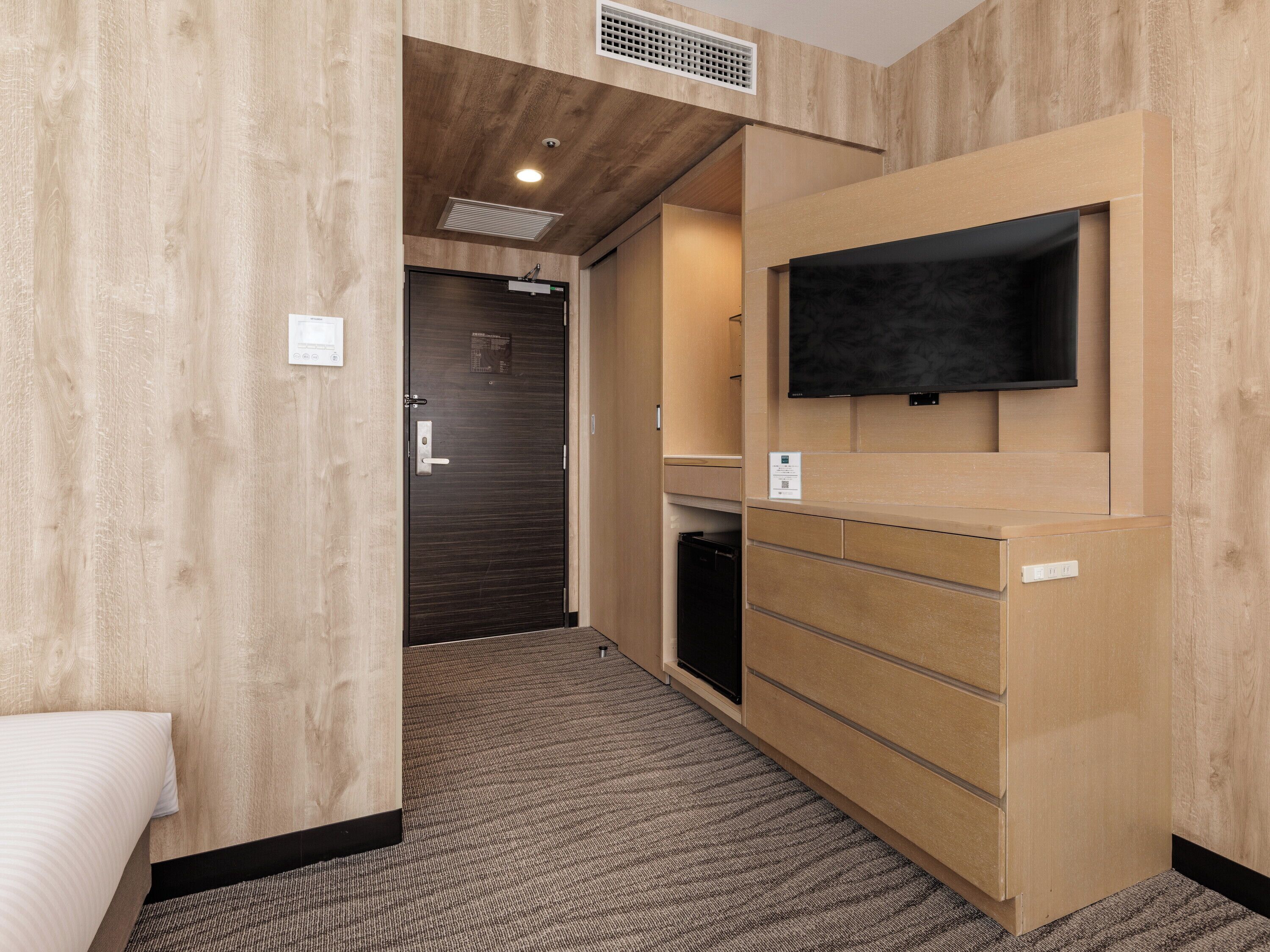 A hotel room with a wooden wall, a flat-screen TV mounted on the wall, a small desk with drawers, and a carpeted floor.