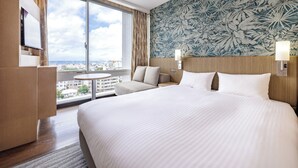 Habitación de hotel con una cama grande, un escritorio, una silla, una mesita y vistas a la ciudad a través de un ventanal amplio.