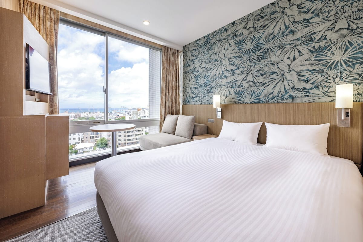 A hotel room with a large bed, a desk, a chair, a small table, and a view of the city through a large window.