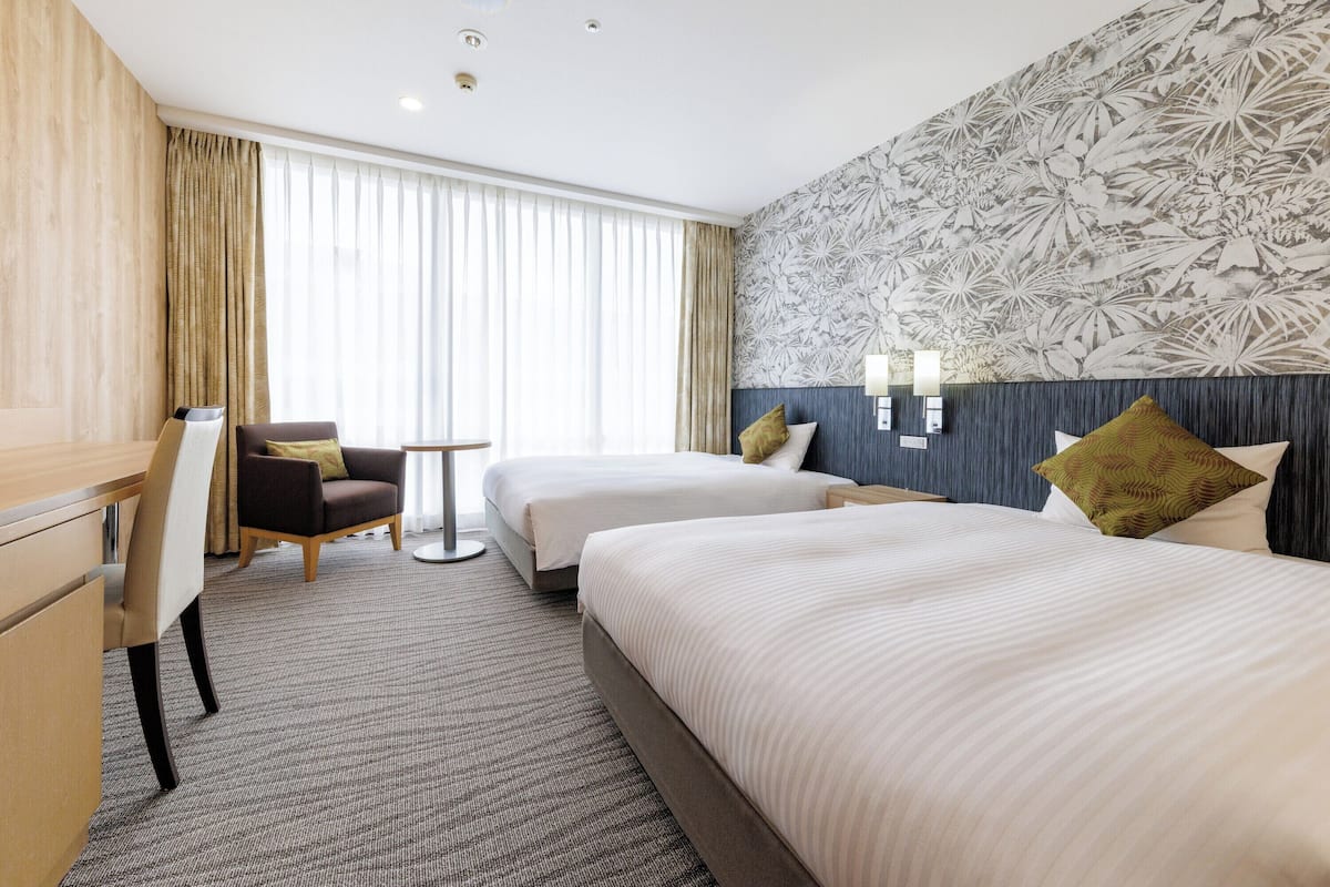 A hotel room with two beds, a desk, a chair, and a large window with curtains.
