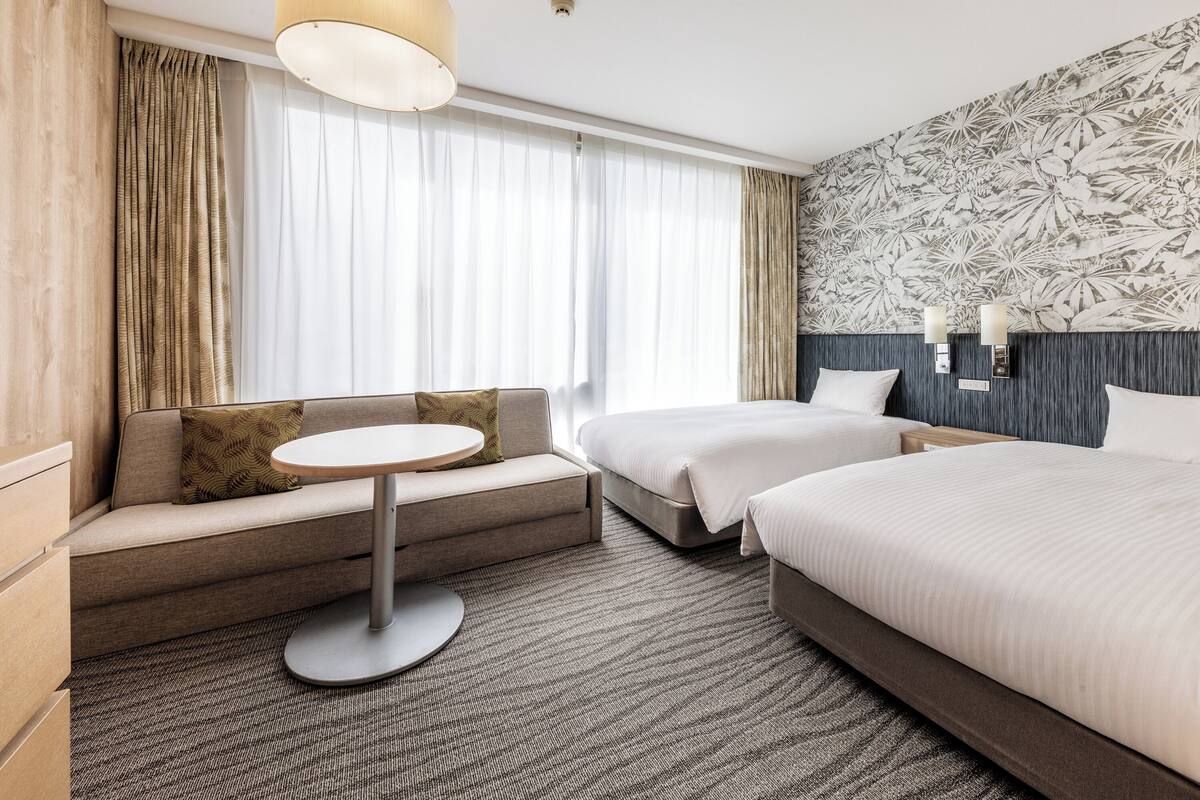 A modern hotel room with a sofa, a round table, two beds, and a patterned wall.