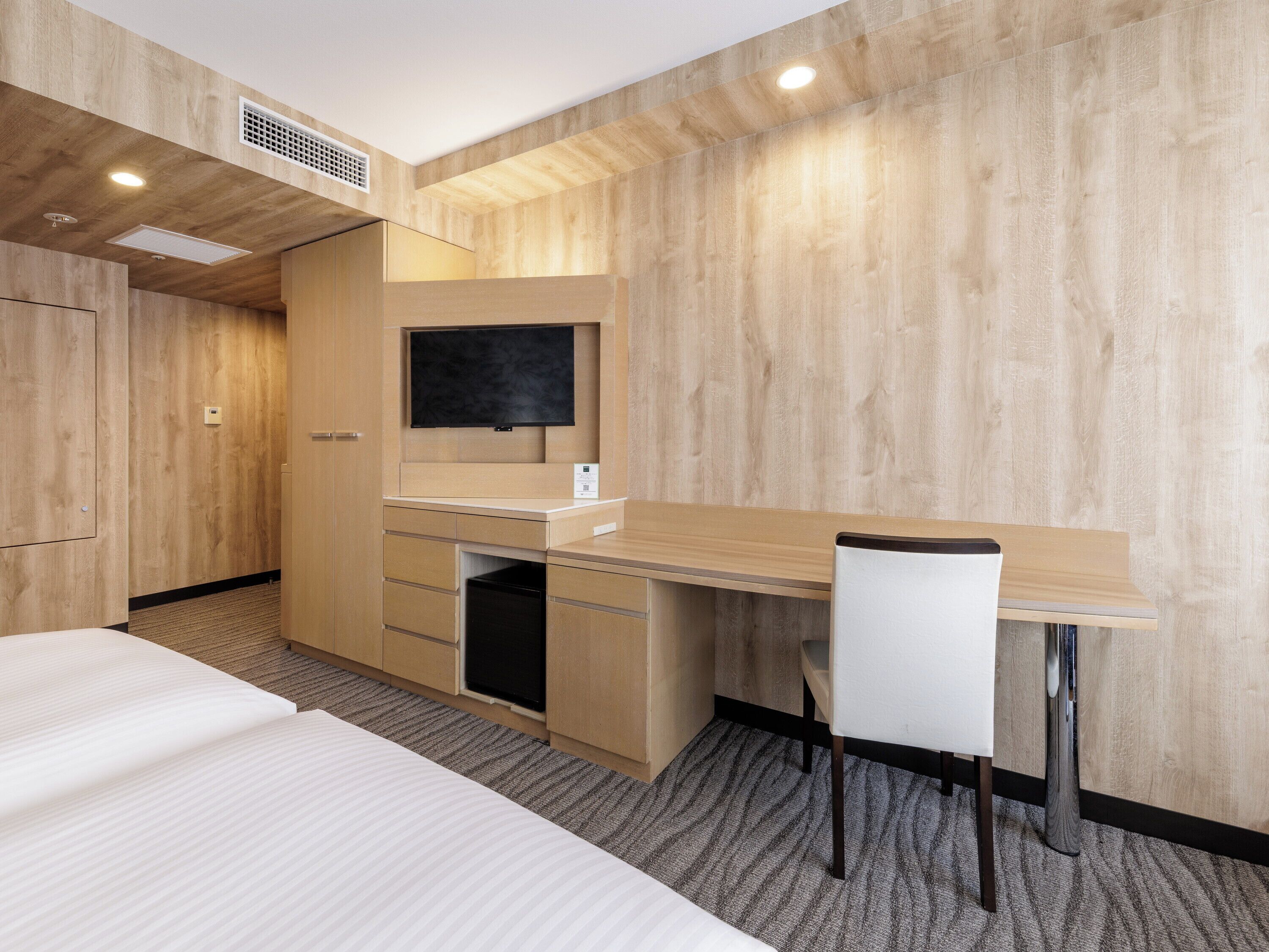A hotel room with a wooden wall, a bed, a desk with a chair, a television, and a built-in heater.