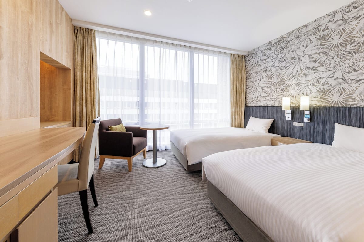A hotel room with two beds, a desk, a chair, and a large window with curtains.