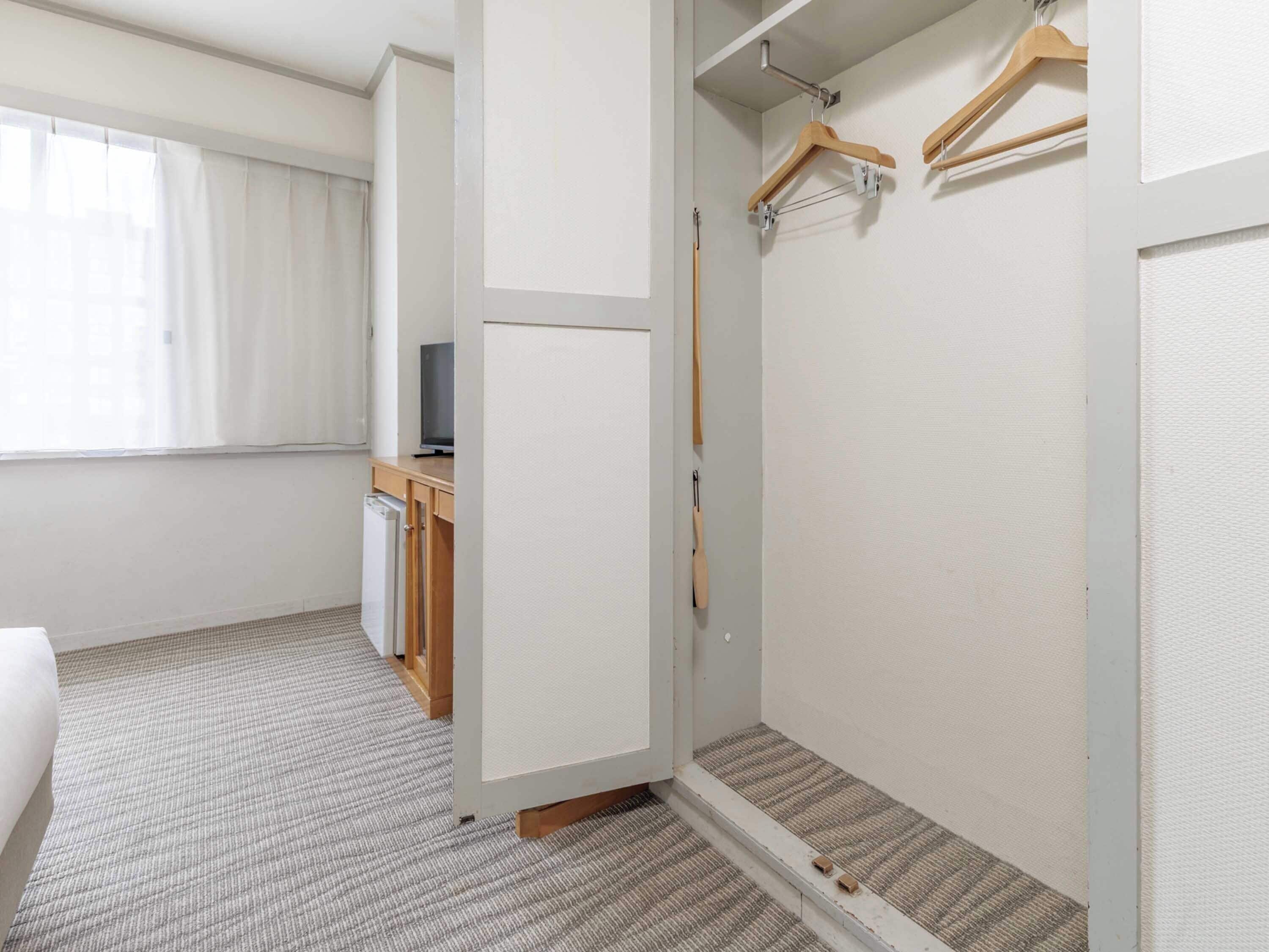 A hotel room with a bed, a desk with a computer, a shower area, and a wardrobe with hangers.