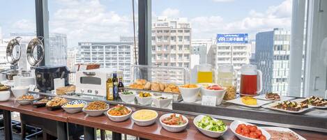 A buffet table with various food items and beverages, set against a cityscape view.
