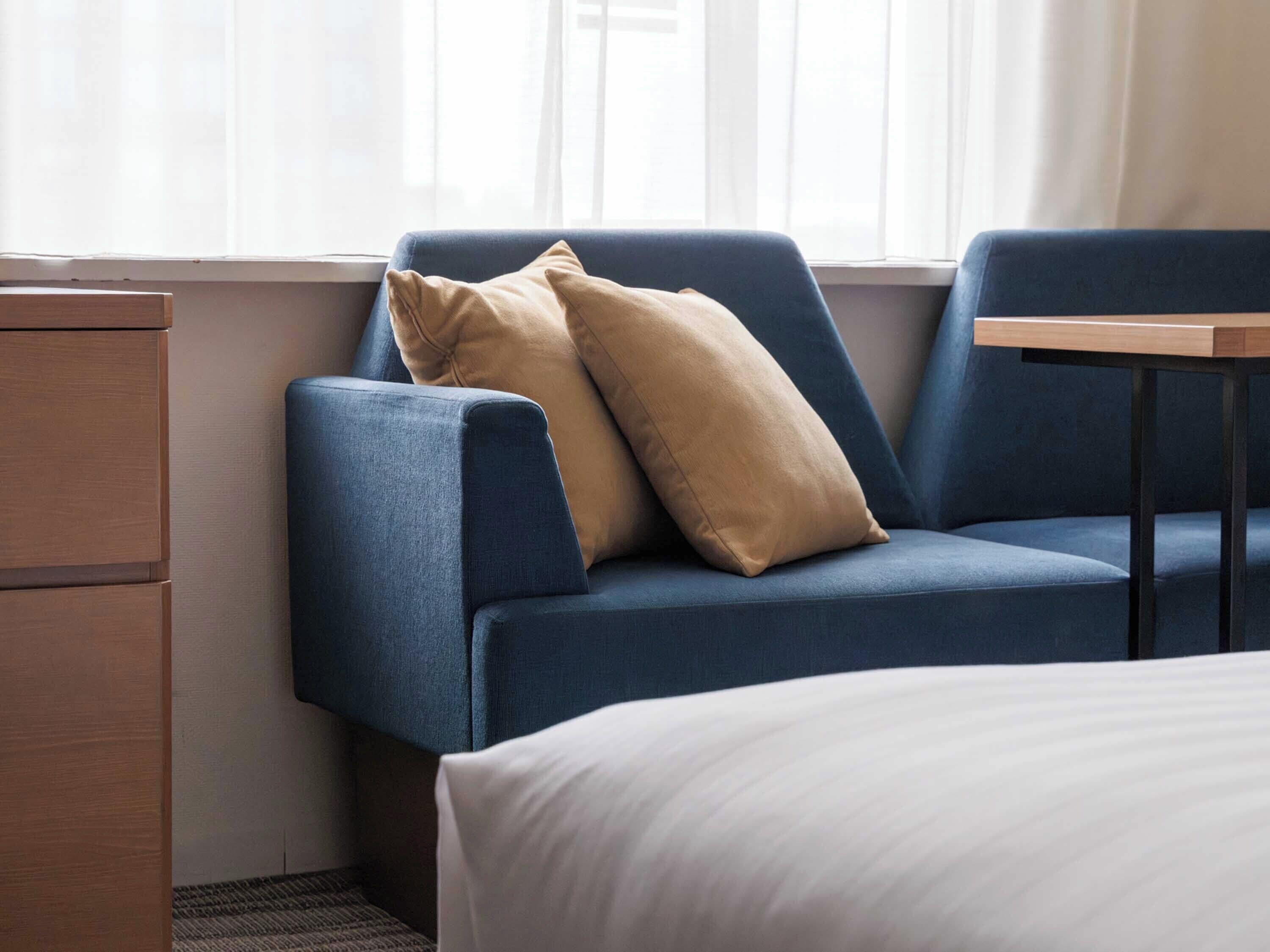 A hotel room with a blue sofa, a wooden nightstand, and a small table.
