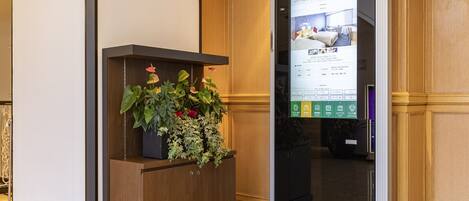 Un espace d’accueil avec un bureau en bois, une plante en pot et un écran numérique affichant des informations sur les chambres.