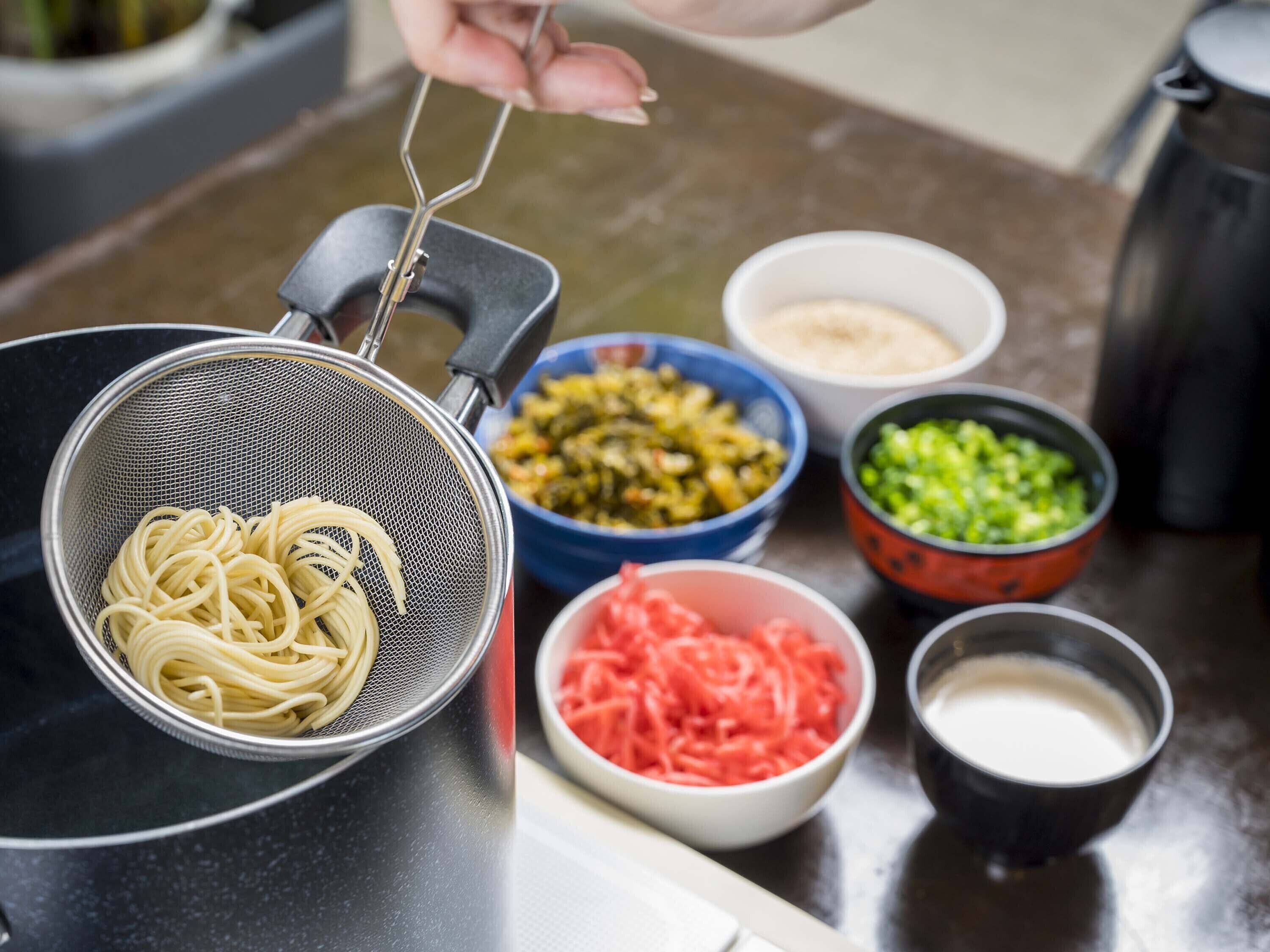 A kitchen with a strainer filled with uncooked pasta, bowls of chopped vegetables, and a pot of milk.