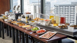 A buffet with various dishes and beverages, a coffee machine, and a view of a cityscape through large windows. - Nishitetsu Inn Fukuoka (Fukuoka)