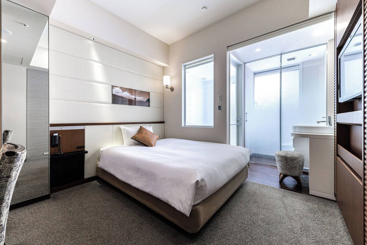 Habitación de hotel moderna con una cama grande, una mesita de noche, un espejo, una ventana y un baño visible a través de una puerta abierta.