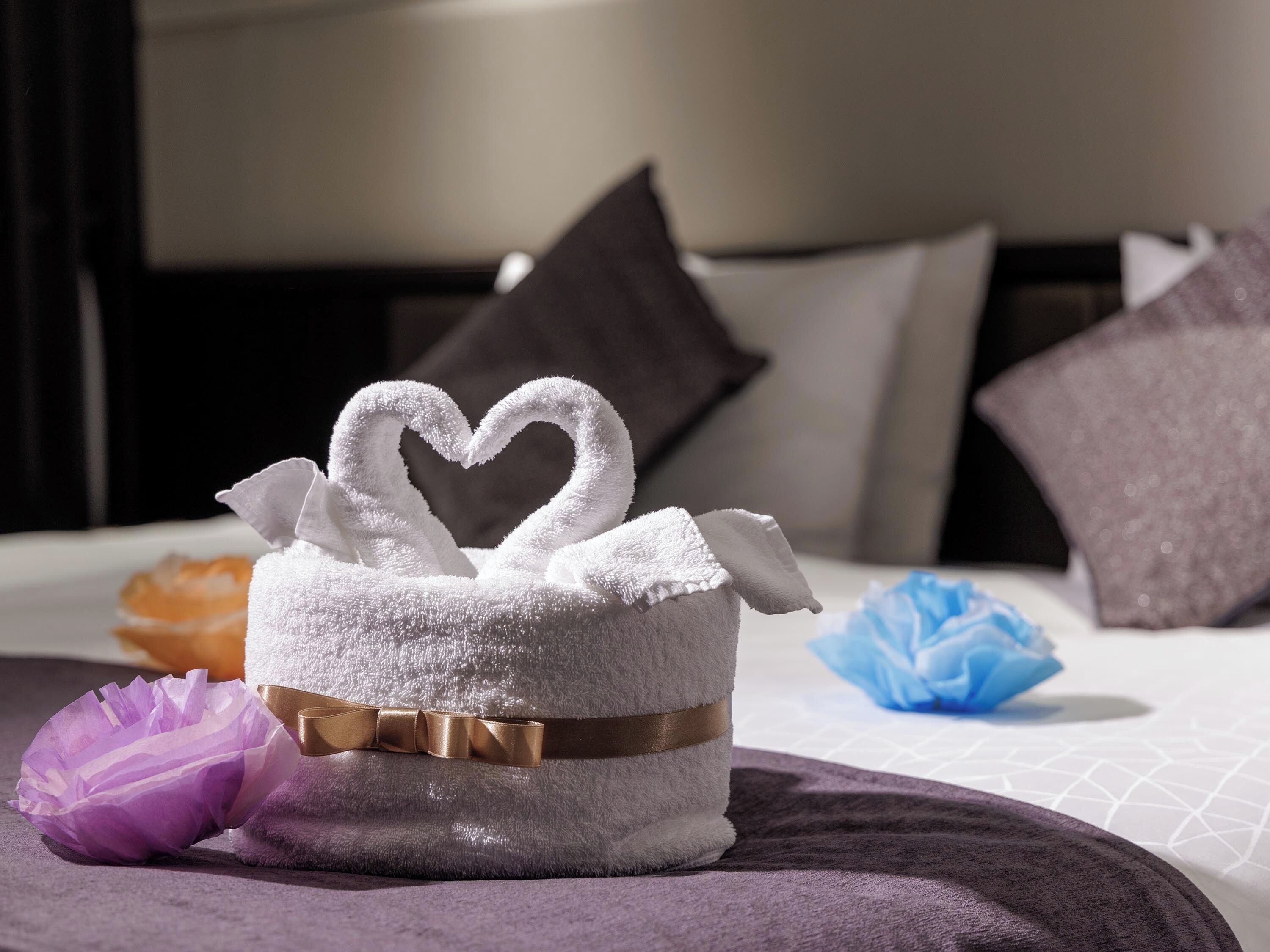 Una toalla doblada con un diseño de corazón, sobre una cama con cubrecama y almohadas moradas.
