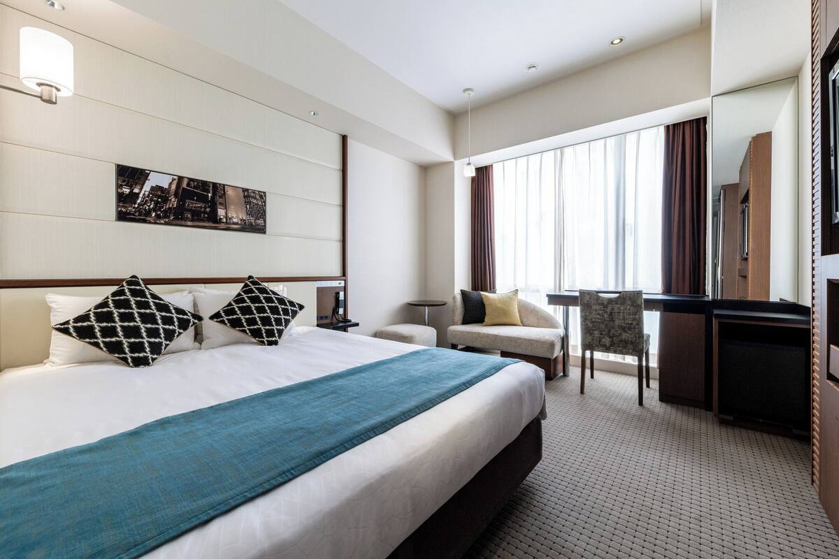 Habitación de hotel con una cama grande, un escritorio, una silla, una mesita y un amplio ventanal con cortinas.