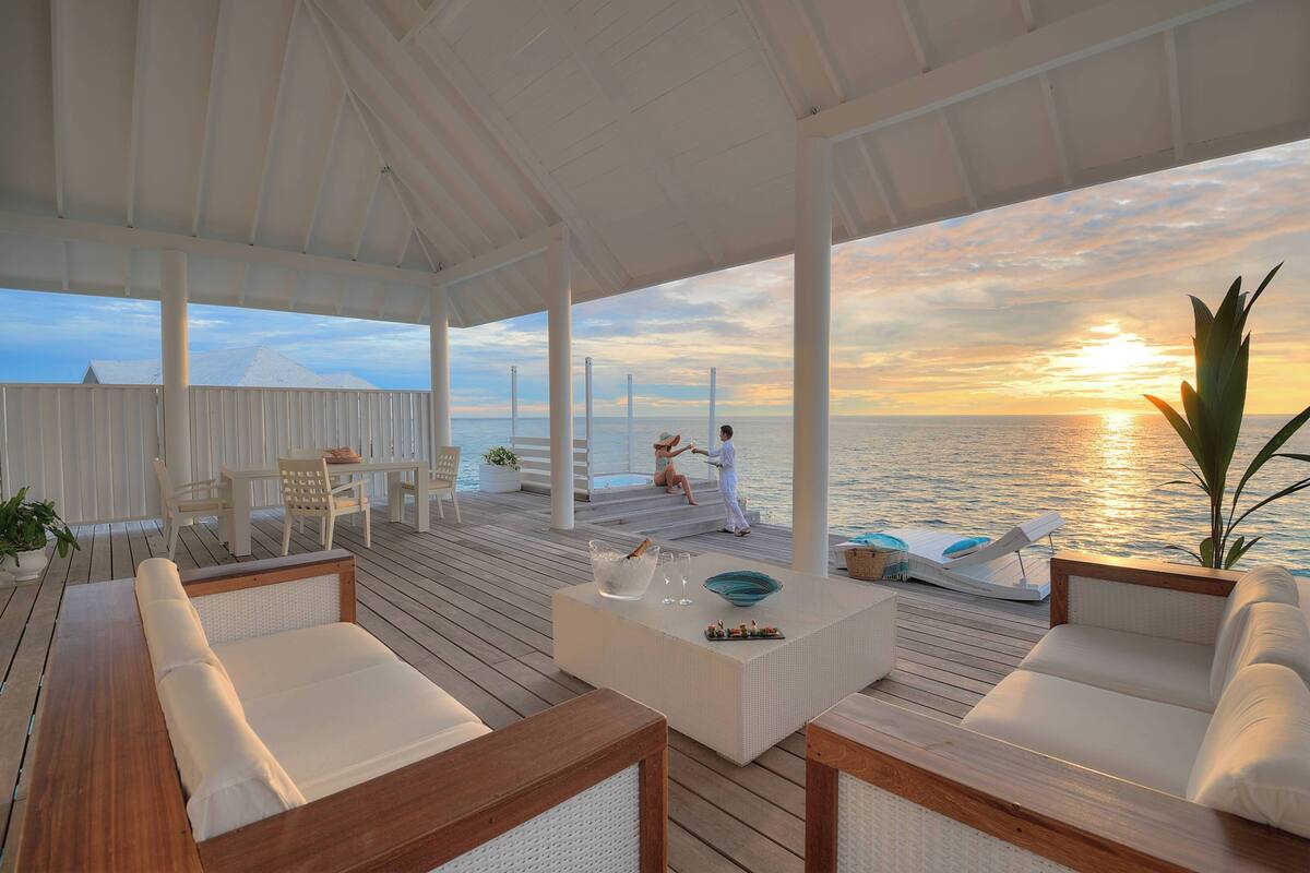 A covered outdoor seating area with a view of the sunset over the water, wooden furniture, and a white table with drinks.
