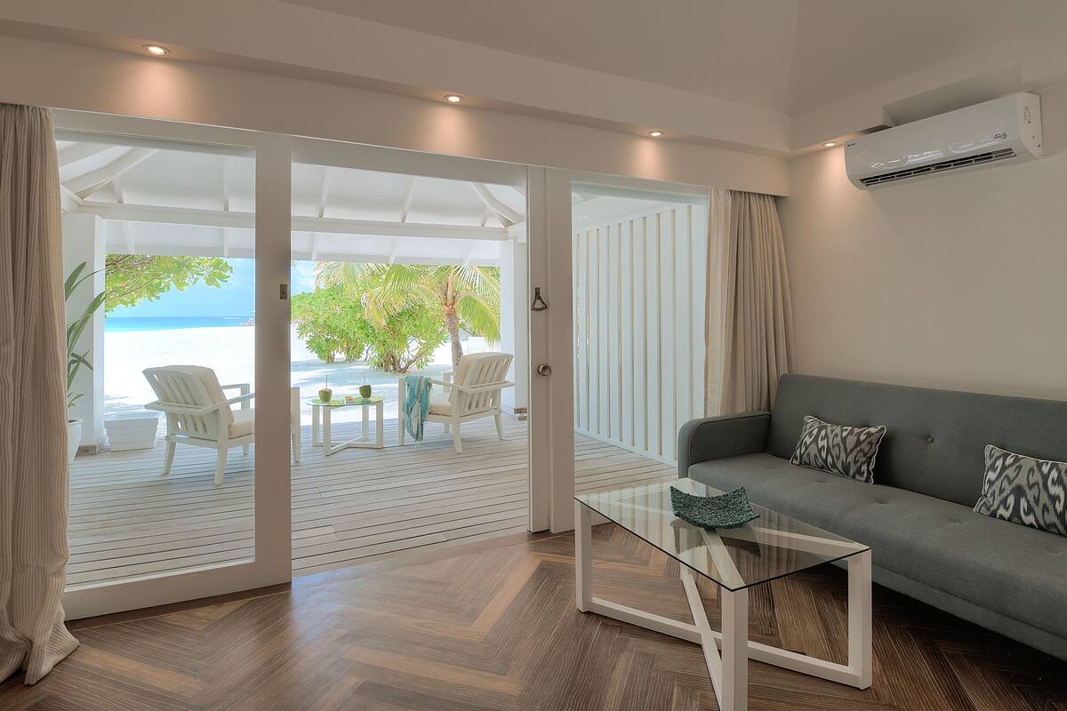 A modern living room with a glass coffee table, a grey sofa, and a view of the beach through large sliding doors.