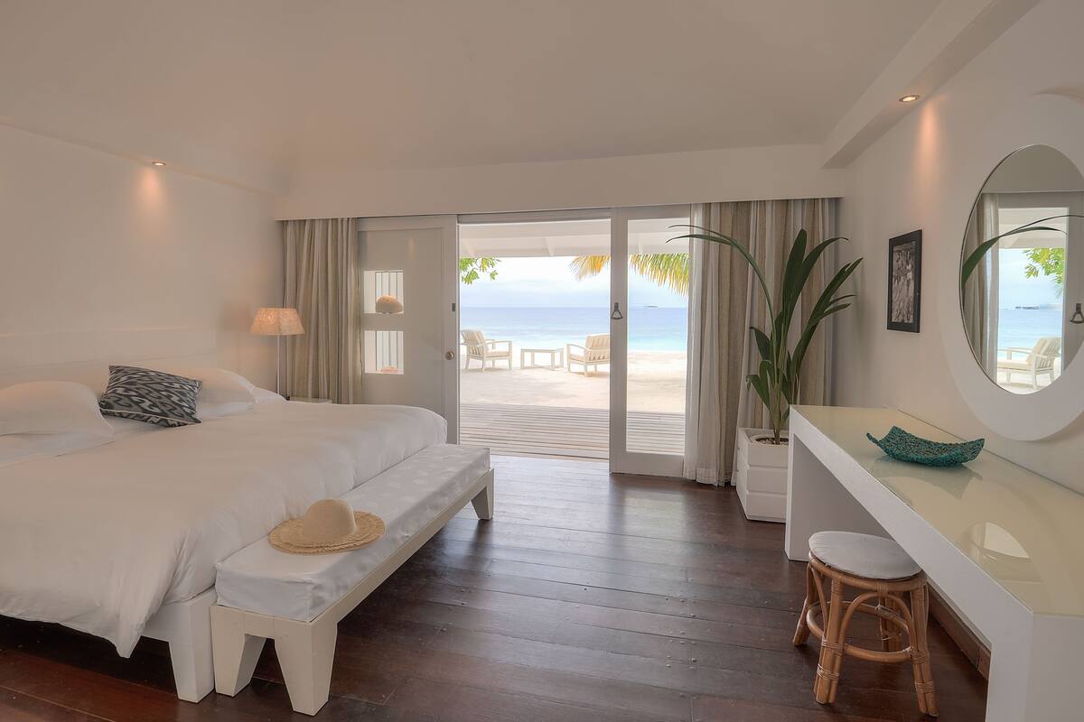 A bedroom with a bed, a bench, a desk, and a view of the beach.
