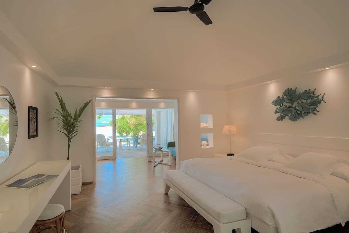A modern bedroom with a large bed, a desk, and a chair. There is a view of a balcony with outdoor furniture.