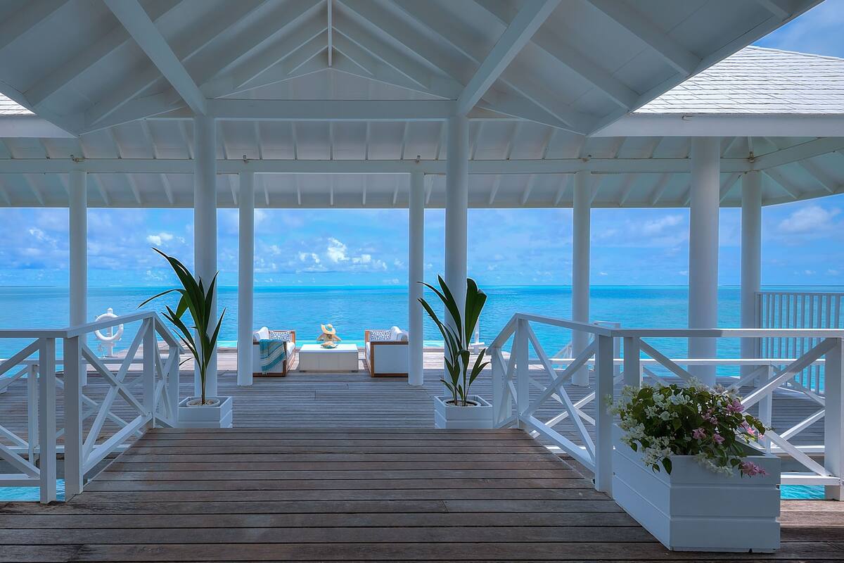 A covered wooden deck with white railings, two potted plants, and a table with chairs facing a clear blue ocean view.