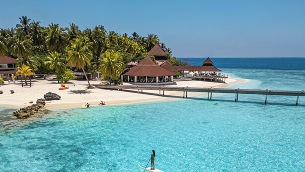 Un océano turquesa transparente con una persona haciendo paddleboard, una playa con palmeras y bungalows sobre el agua.