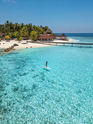 A clear turquoise ocean with a person paddleboarding, a beach with palm trees, and overwater bungalows.