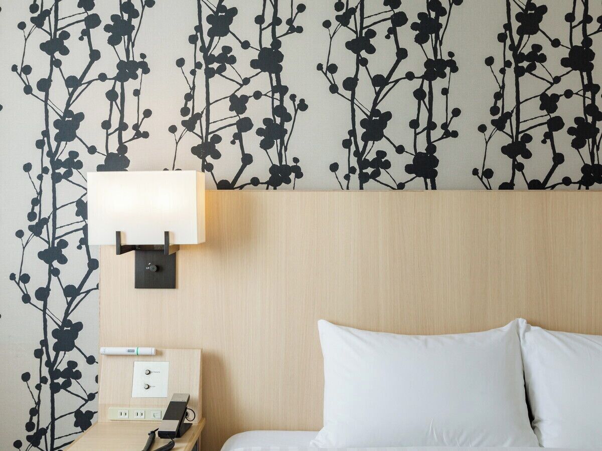 A hotel room with a bed, a nightstand, a lamp, and a wall with a black floral pattern.