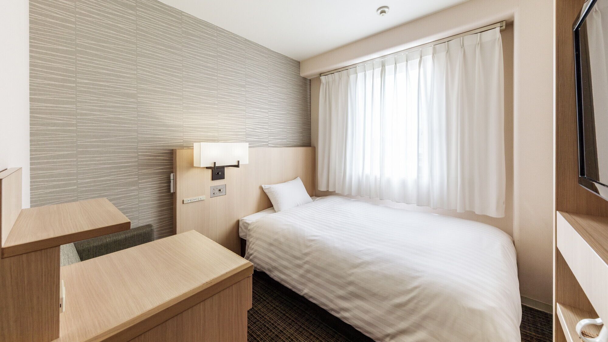 A hotel room with a bed, a desk, a wall-mounted lamp, and a window with curtains.