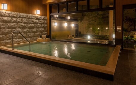 A spa pool with a stone wall, wooden ceiling, and a view of a waterfall through a glass wall.