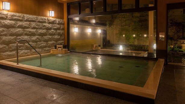 Un bassin de spa avec un mur en pierre, un plafond en bois et une vue sur une cascade, visible à travers une paroi en verre.