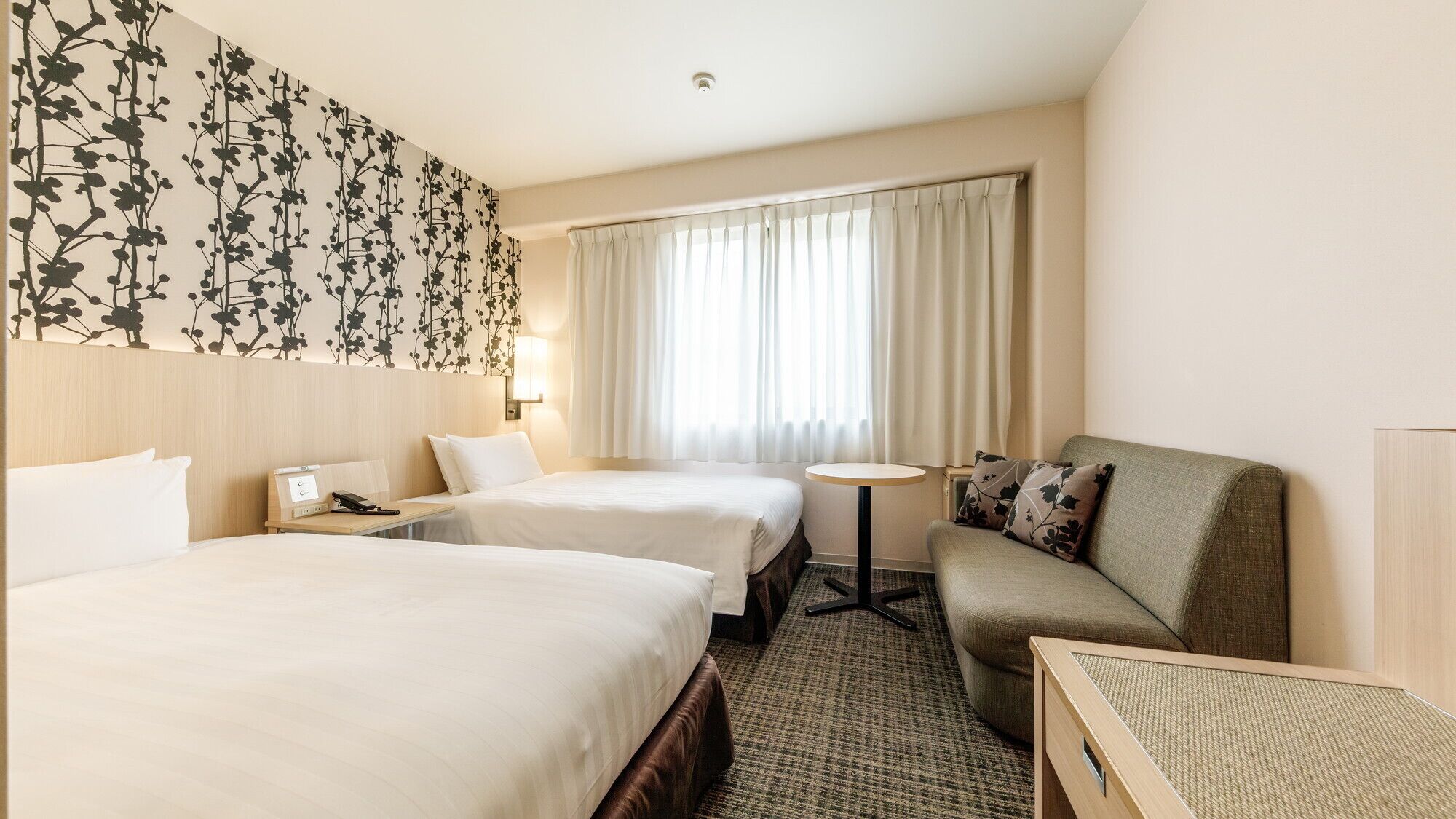 A hotel room with two beds, a sofa, a small table, a nightstand, and a wall with a floral pattern.