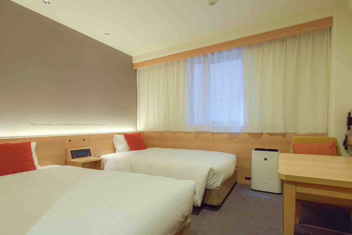 A hotel room with two beds, a desk, a chair, a bedside table, and a window with curtains.