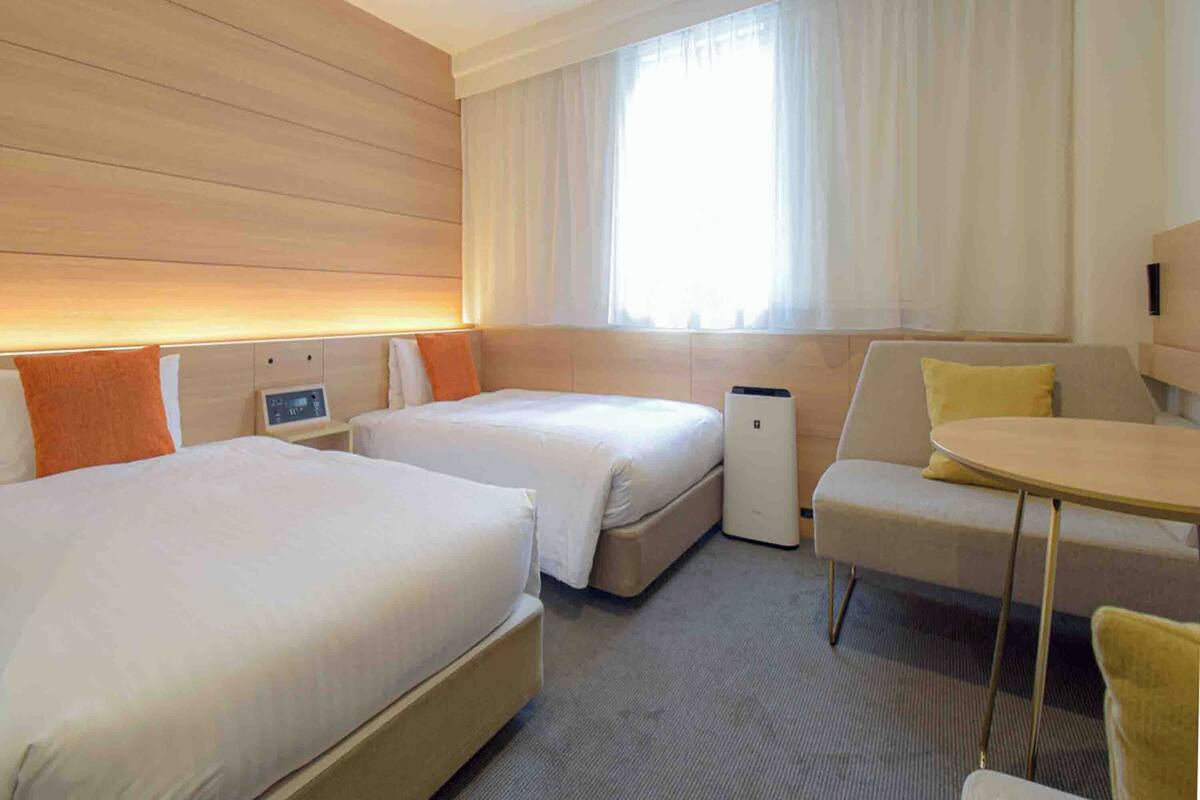 A hotel room with two beds, a small table, a sofa, and a window with curtains.