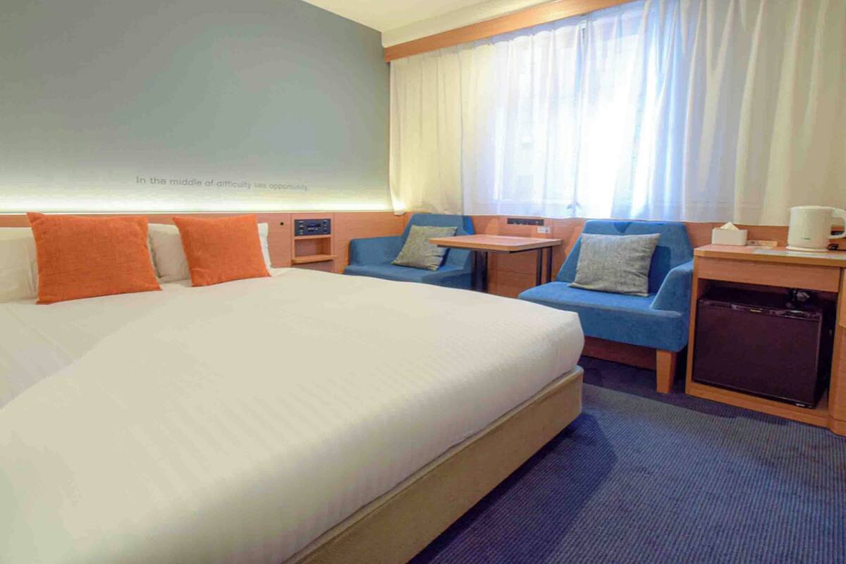 A hotel room with a large bed, a small table, two blue armchairs, a desk with a computer, and a window with curtains.