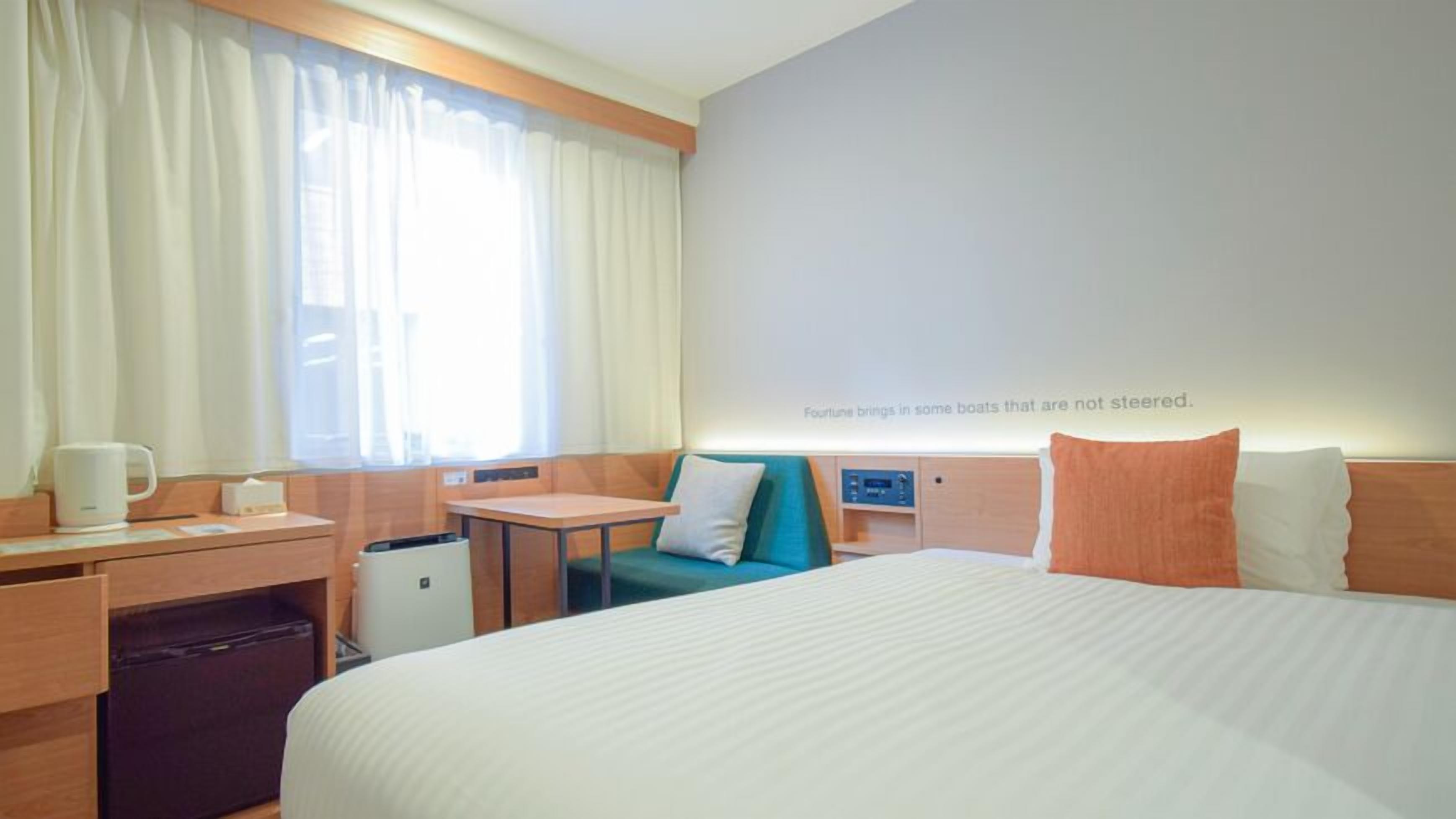 Hotellihuone, jossa on sänky, työpöytä, tuoli, pieni pöytä, roskakori ja ikkuna, jossa on verhot.